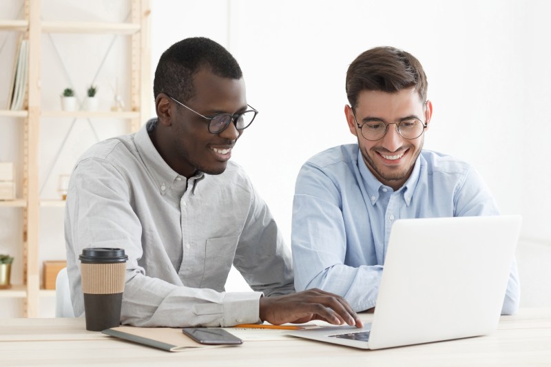 Executive manager or startup mentor explaining work to young focused intern, looking at laptop screen in white office, training new worker